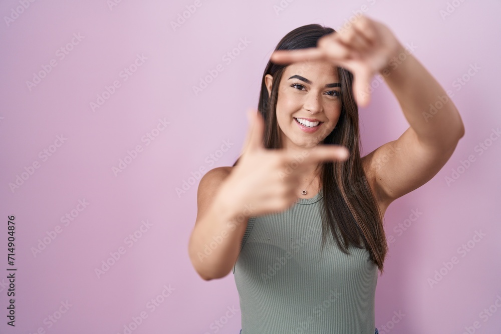 Fototapeta premium Hispanic woman standing over pink background smiling making frame with hands and fingers with happy face. creativity and photography concept.