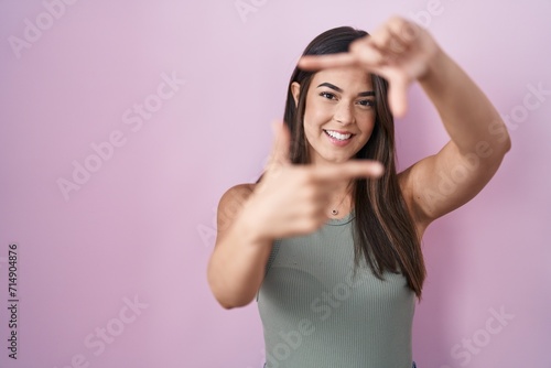 Hispanic woman standing over pink background smiling making frame with hands and fingers with happy face. creativity and photography concept.