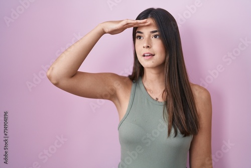 Hispanic woman standing over pink background very happy and smiling looking far away with hand over head. searching concept.