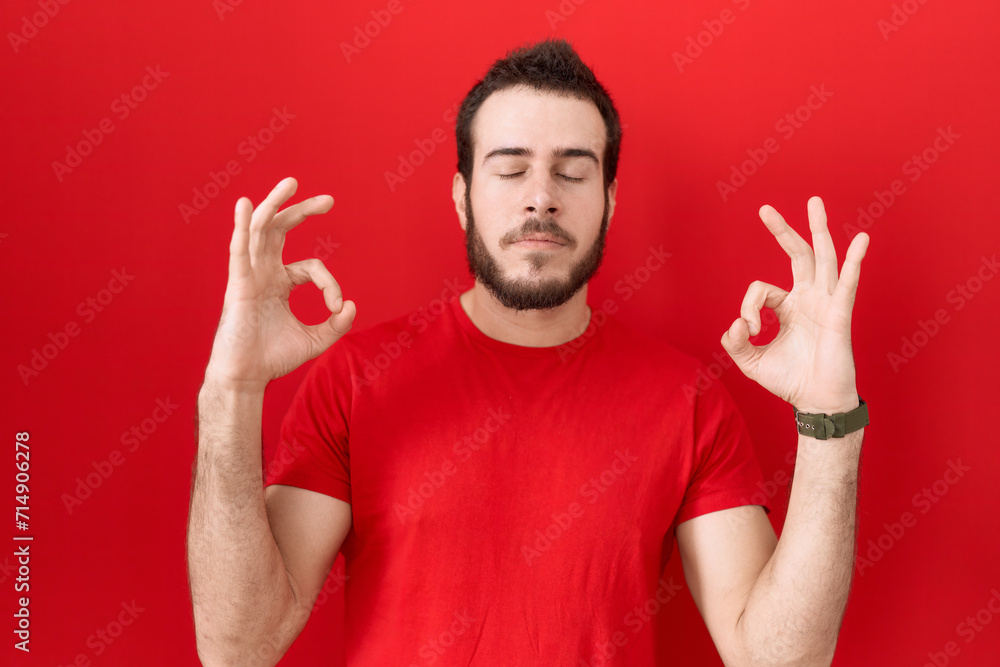 Young hispanic man wearing casual red t shirt relaxed and smiling with eyes closed doing meditation gesture with fingers. yoga concept. Young hispanic man wearing casual red t shirt relaxed and smiling with eyes closed doing meditation gesture with fingers. yoga concept.