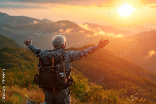 Close up back view of old elderly man with a travel backpack on his back and holds his hands up as a sign of freedom standing on rock looking at mountains. Joyful free travel concept