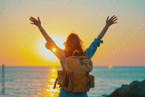 Close up back view of young woman with a travel backpack on her back and holds her hands up as a sign of freedom standing on rock looking at sea ocean. Joyful free travel concept photo