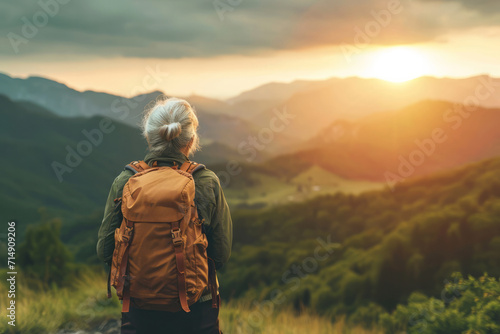 Close up back view of a calm elderly old mature woman in a travel backpack standing on rock looking at mountains. Travelling concept.