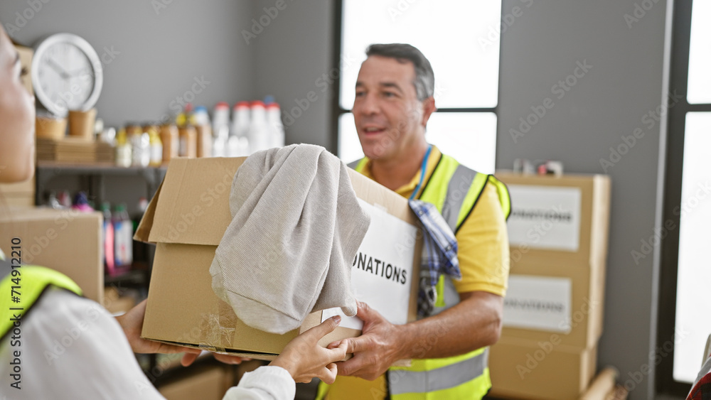 Cheerful man and woman volunteers confidently giving cardboard box full of clothes donation at buzzing charity center Cheerful man and woman volunteers confidently giving cardboard box full of clothes donation at buzzing charity center