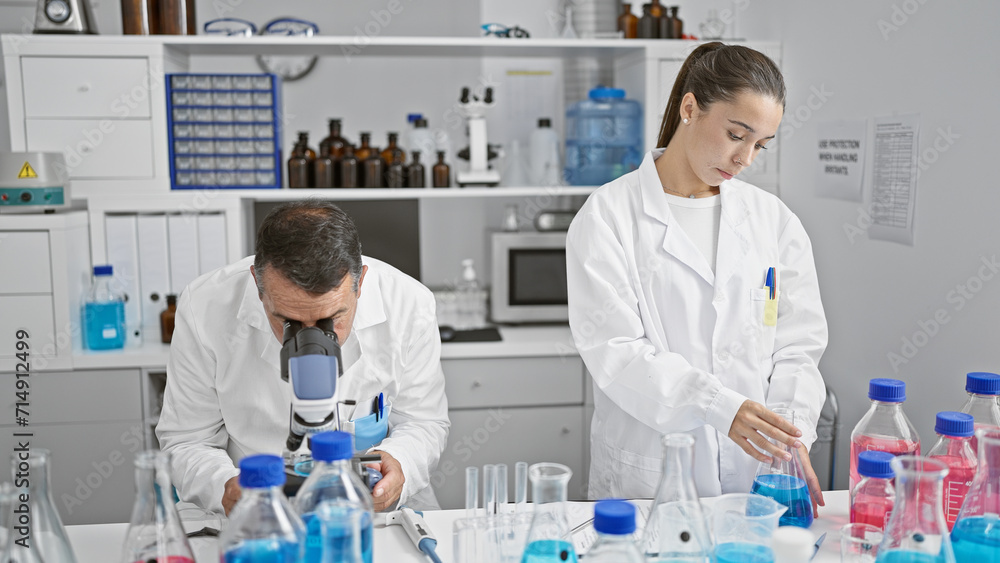 Hispanic man and woman scientists working together, engrossed in medical research at their indoor laboratory, measuring liquid samples while peering through their microscope. Hispanic man and woman scientists working together, engrossed in medical research at their indoor laboratory, measuring liquid samples while peering through their microscope.