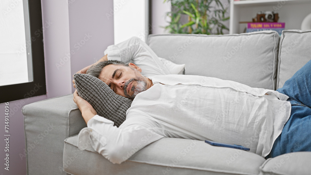 Exhausted young hispanic man with handsome grey-haired beard comfortably lying on the cozy sofa, deeply sleeping and enjoying relaxation in the living room of his home, escaping the tired reality. Exhausted young hispanic man with handsome grey-haired beard comfortably lying on the cozy sofa, deeply sleeping and enjoying relaxation in the living room of his home, escaping the tired reality.