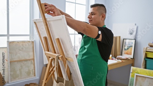 Young, confident latin artist joyfully standing by his easel in the art studio, ready to transfer creativity onto canvas