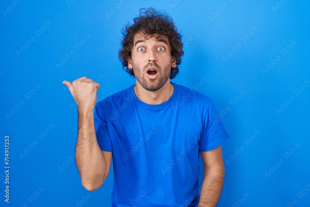 Naklejka premium Hispanic young man standing over blue background surprised pointing with hand finger to the side, open mouth amazed expression.