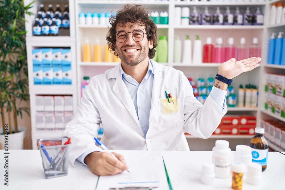 Hispanic young man working at pharmacy drugstore smiling cheerful presenting and pointing with palm of hand looking at the camera.