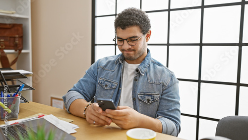 A young man with glasses and a beard uses a smartphone in a modern office setting.