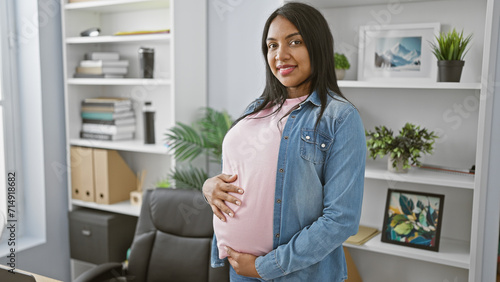 Smiling young pregnant woman confidently touches belly while working in the office, embodying maternity and business success.