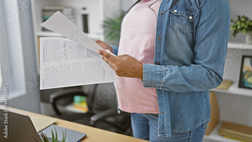Expecting young worker, pregnant businesswoman reading documents, tenderly touching her belly, standing indoors at the office photo