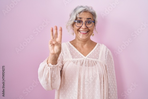 Slika na platnu Middle age woman with grey hair standing over pink background showing and pointing up with fingers number three while smiling confident and happy