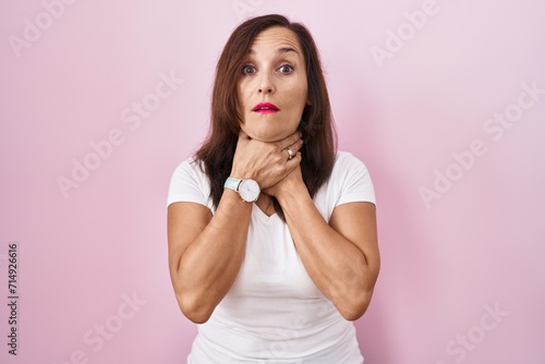 Middle age brunette woman standing over pink background shouting suffocate because painful strangle. health problem. asphyxiate and suicide concept.