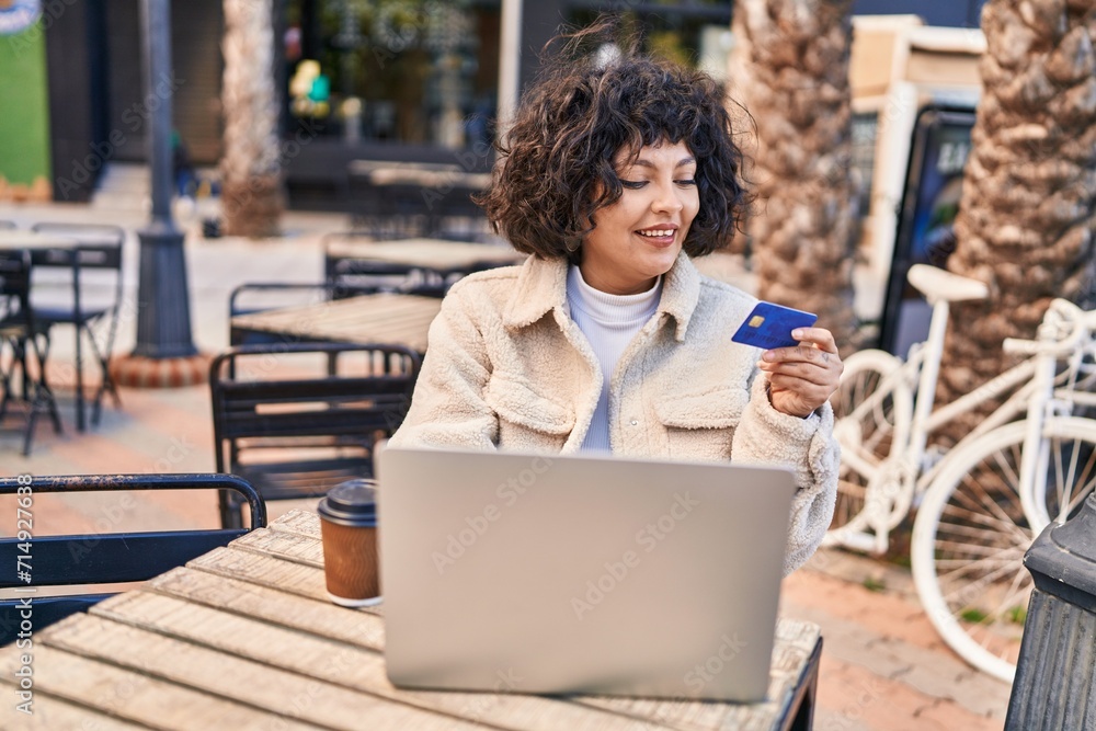 Young beautiful hispanic woman using laptop and credit card sitting on table at coffee shop terrace