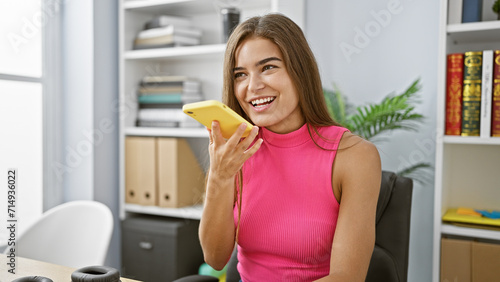 Empowered young hispanic business woman confidently sends voice message from her smartphone  radiating success while working in her sleek office