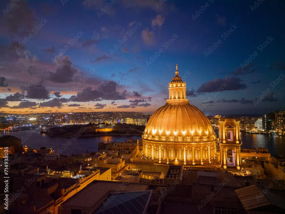Aerial view of illuminated dome of Basilica of Our Lady of Mount Carmel in Valletta, Malta. View towards Manoel island with fort and Sliema skyline.