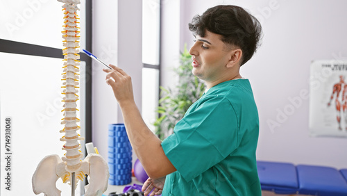 A young man in scrubs examines a spinal model in a brightly lit medical clinic's interior. photo