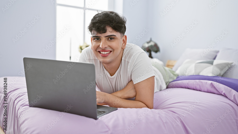 A smiling man lying on a purple bed using a laptop in a bright bedroom A smiling man lying on a purple bed using a laptop in a bright bedroom
