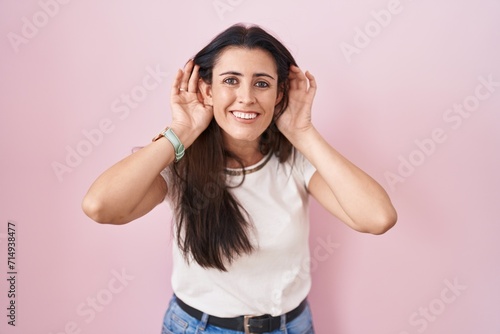 Young brunette woman standing over pink background trying to hear both hands on ear gesture, curious for gossip. hearing problem, deaf