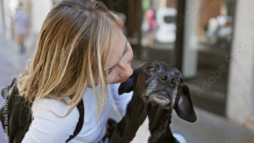 A young blonde woman lovingly gazes at her black labrador on a sunlit urban street.