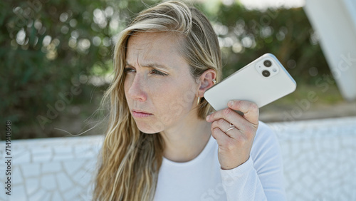 Fotografie Young blonde woman listening to a voicemail outdoors in a green park, with a con