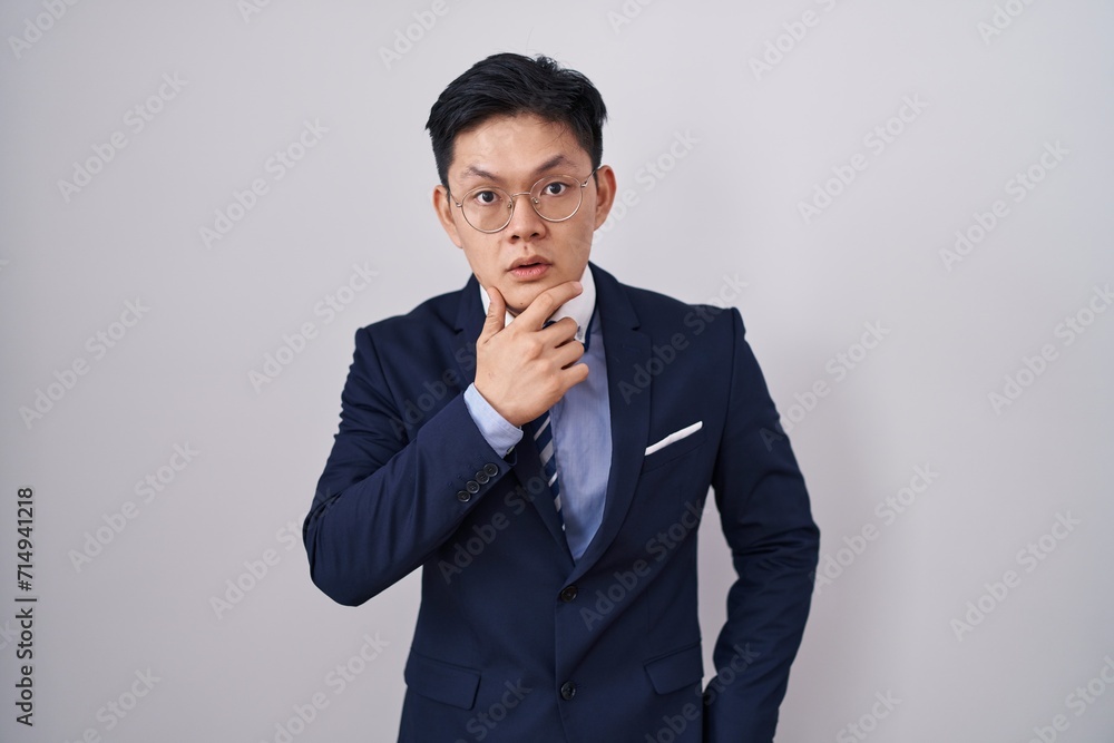 Young asian man wearing business suit and tie looking fascinated with disbelief, surprise and amazed expression with hands on chin Young asian man wearing business suit and tie looking fascinated with disbelief, surprise and amazed expression with hands on chin