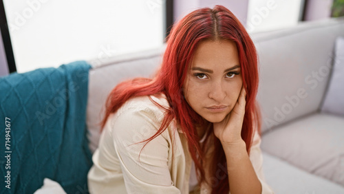Worried redhead adult sitting on the home sofa caught in a web of doubt, a sad expression painting the beautiful young woman's portrait in her living room