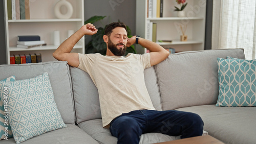 Young hispanic man sitting on sofa tired stretching arms at home