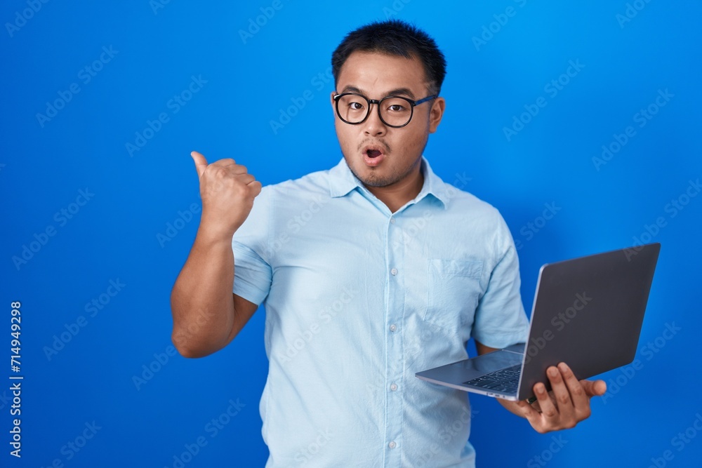 Chinese young man using computer laptop surprised pointing with hand finger to the side, open mouth amazed expression. Chinese young man using computer laptop surprised pointing with hand finger to the side, open mouth amazed expression.