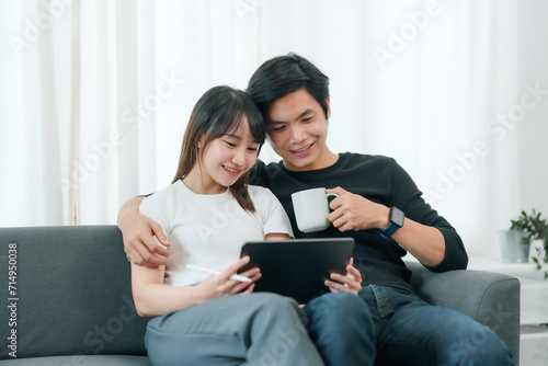 Young asian couple sitting on couch, happily sharing a moment with a tablet, conveying leisure and technology in daily life..