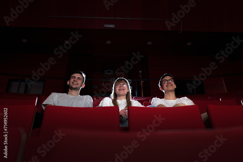Three friends, a teenage girl and two boys smiling, watching a movie in an empty cinema with red chairs. Young adults and leisure concepts.