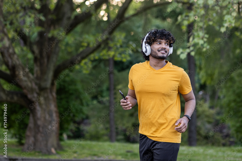 Young smiling Indian man running in the park wearing headphones and holding a mobile phone