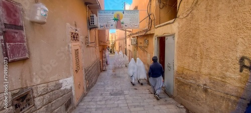 narrow alley, people wearing traditional clothes, and houses made of stones and clay, typical sub-Saharan architecture of the city of Ghardaïa. Oasis M'zab, Algeria. photo