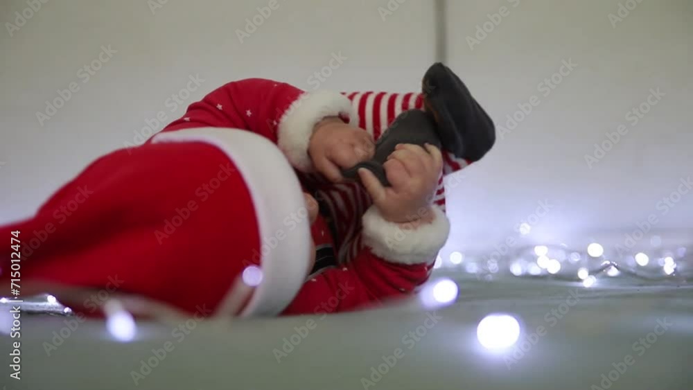A little boy in a santa claus costume with a red hat pulls up his leg to his face, tries to gnaw it, plays with his leg against the background of lights for New Year and Christmas A little boy in a santa claus costume with a red hat pulls up his leg to his face, tries to gnaw it, plays with his leg against the background of lights for New Year and Christmas