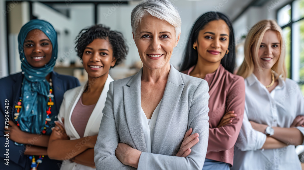 Group of diverse professional women confidently standing in a line, with the woman in the foreground crossing her arms, showcasing a strong and united front in a workplace setting. Group of diverse professional women confidently standing in a line, with the woman in the foreground crossing her arms, showcasing a strong and united front in a workplace setting.