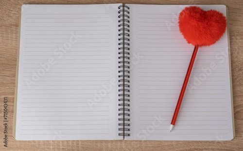 A notebook lies on a wooden table. An empty notebook is open. A red heart on a pen lies on a notebook page.