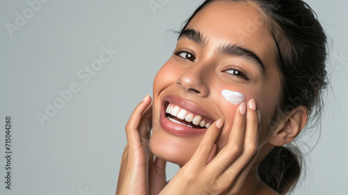 Close-up of a woman with clear blue eyes and flawless skin applying a white cream on her cheek with her fingers, exemplifying a skincare routine.