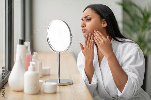 Upset young indian woman looking at mirror, touching her face photo
