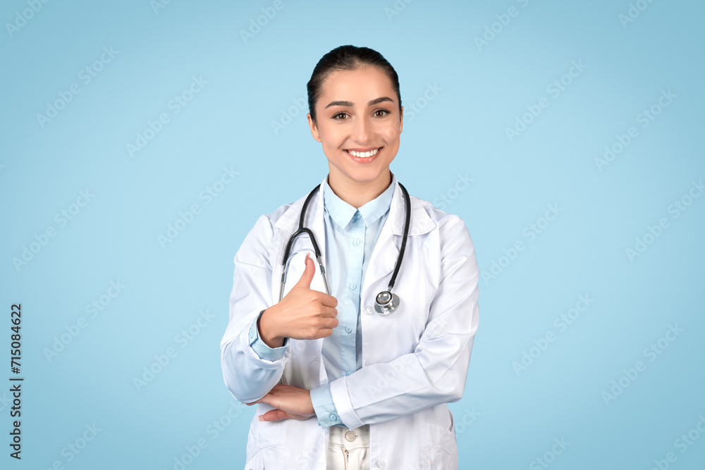 Happy young female doctor with stethoscope around her neck giving thumbs up and smiling, blue backdrop Happy young female doctor with stethoscope around her neck giving thumbs up and smiling, blue backdrop