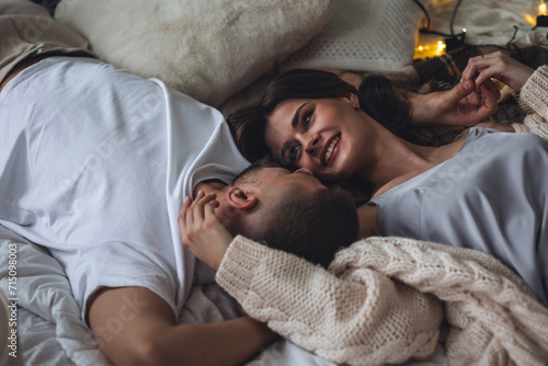 Beautiful happy loving young smiling couple relaxing in bed, looking at each other. Cozy home atmosphere, tenderness, closeness. Embracing, kissing. Lazy weekend, slow living concept