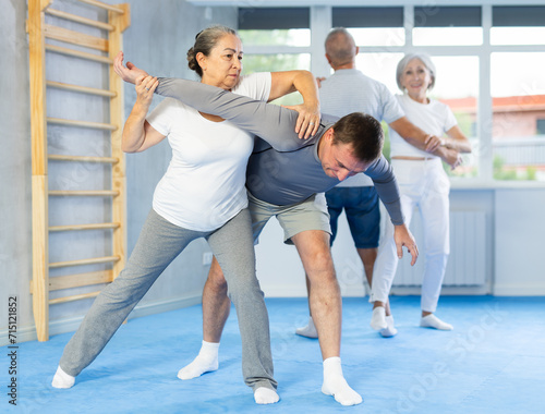 Elderly woman twists the arm of attacking man with painful hold in gym. Self-defense lesson