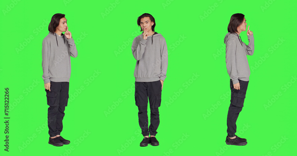 Obraz premium Adult doing hush secret sign over greenscreen, expressing secrecy symbol with finger over lips. Young person showing mute silence gesture , being private in studio with full body backdrop.