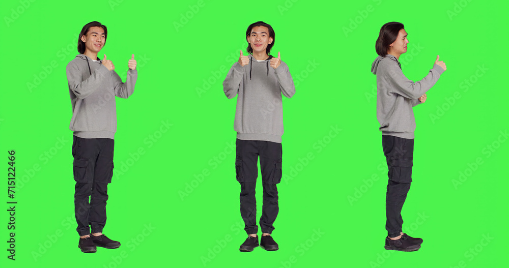 Obraz premium Asian guy does thumbs up gesture standing over full body greenscreen, showing agreement and presenting like symbol in studio. Young joyful adult feeling pleased, approval sign.