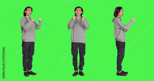 Asian guy does thumbs up gesture standing over full body greenscreen  showing agreement and presenting like symbol in studio. Young joyful adult feeling pleased  approval sign.