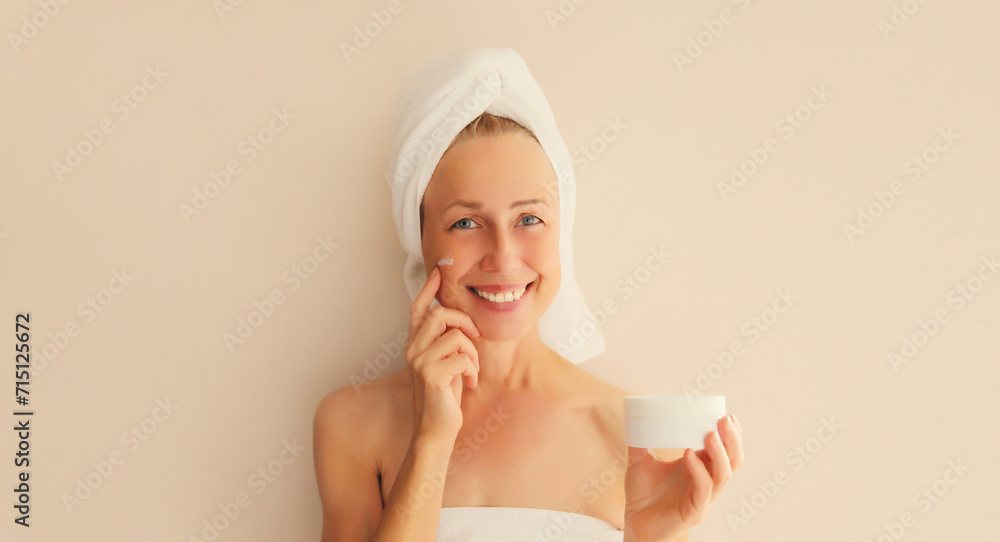 Natural beauty portrait of happy smiling caucasian middle-aged woman touches her clean skin applying face cream while drying wet hair with white wrapped bath towel on her head after shower in morning Natural beauty portrait of happy smiling caucasian middle-aged woman touches her clean skin applying face cream while drying wet hair with white wrapped bath towel on her head after shower in morning