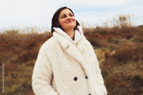 Beautiful young brunette swarthy woman in white coat in nature in fall, winter cold day. Female smile, happiness, well-being. Happy girl outdoors.