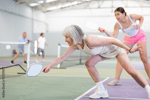 Focused active aged woman playing friendly pickleball match on indoor court. Senior people sports concept..