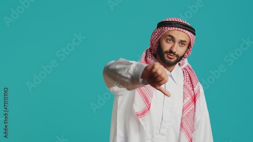 Obraz na plátně Muslim guy with ancient gown and kufiyah gives thumbs down and feels unsatisfied with stuff