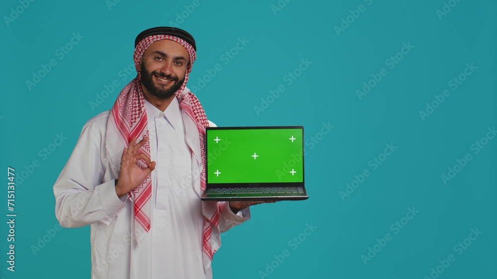 Muslim adult shows greenscreen laptop with blank mockup layout on display, copyspace template. Arab guy wearing gown and headscarf, presenting isolated chromakey screen on pc. Muslim adult shows greenscreen laptop with blank mockup layout on display, copyspace template. Arab guy wearing gown and headscarf, presenting isolated chromakey screen on pc.